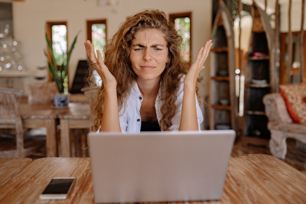 mulher de cabelo ondulado castanho claro sentada em frente a um laptop com as duas mãos levantadas na altura do ombro e expressão confusa