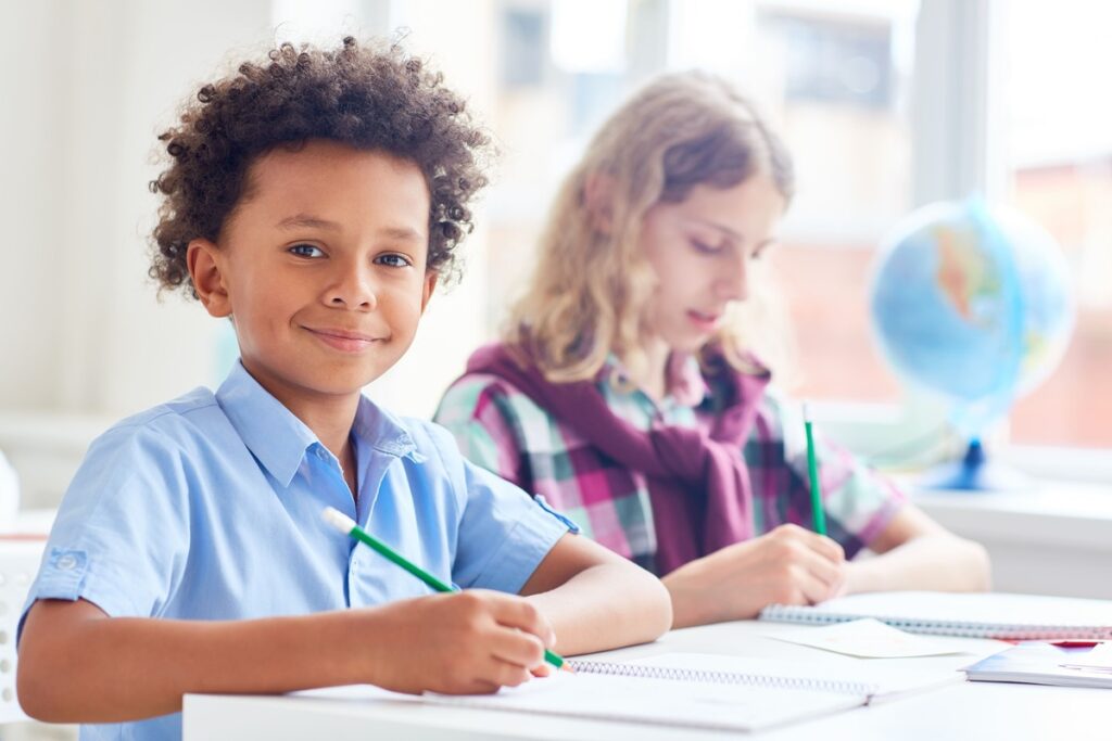 criança negra sentada com lápis na mão. Ao seu lado há uma criança branca sentada, escrevendo