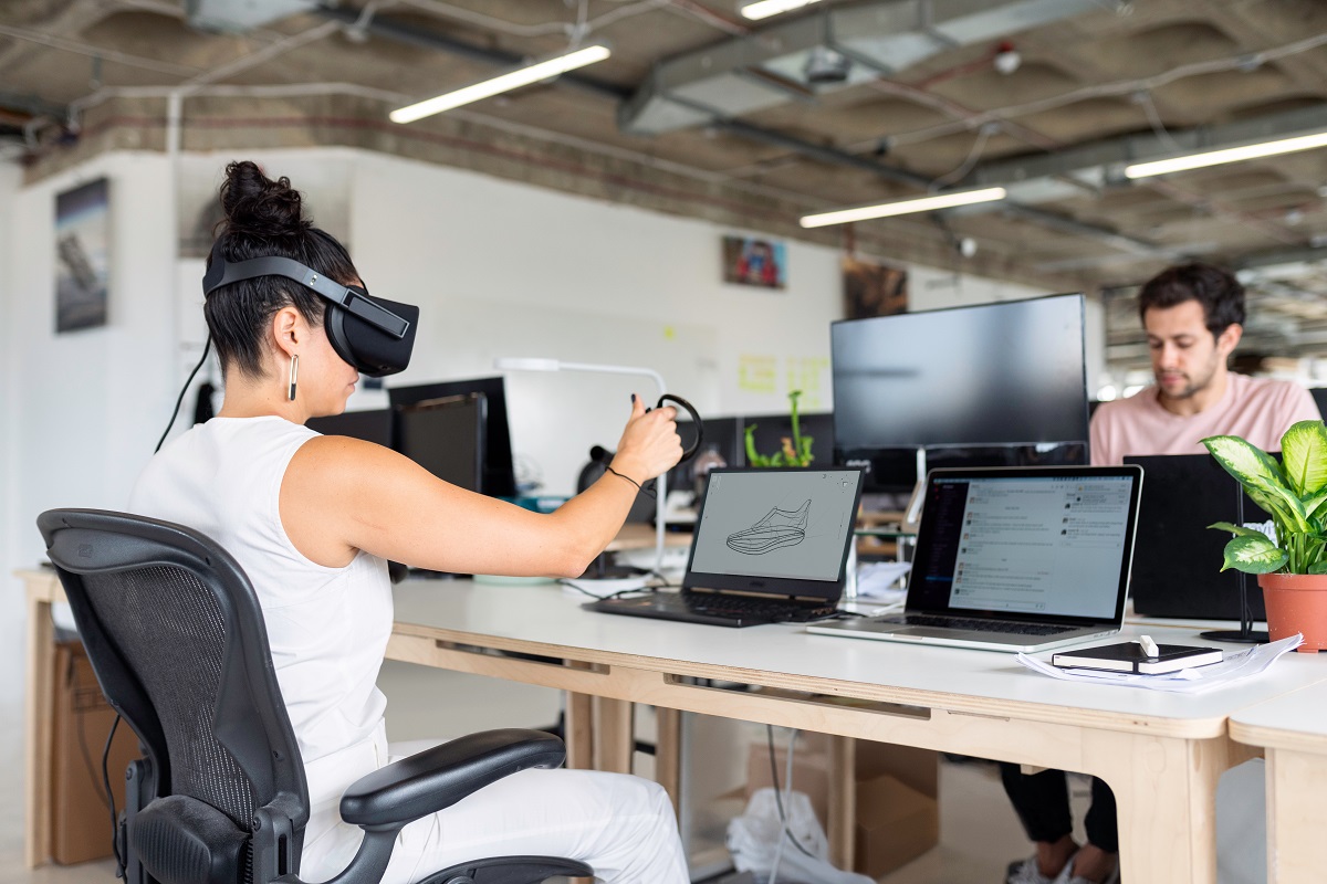 Uma pessoa usando um óculos de realidade virtual, sentada em frente ao computador e fazerndo um projeto de design. Na sua frente, outro colaboraor trabalha ao computador.