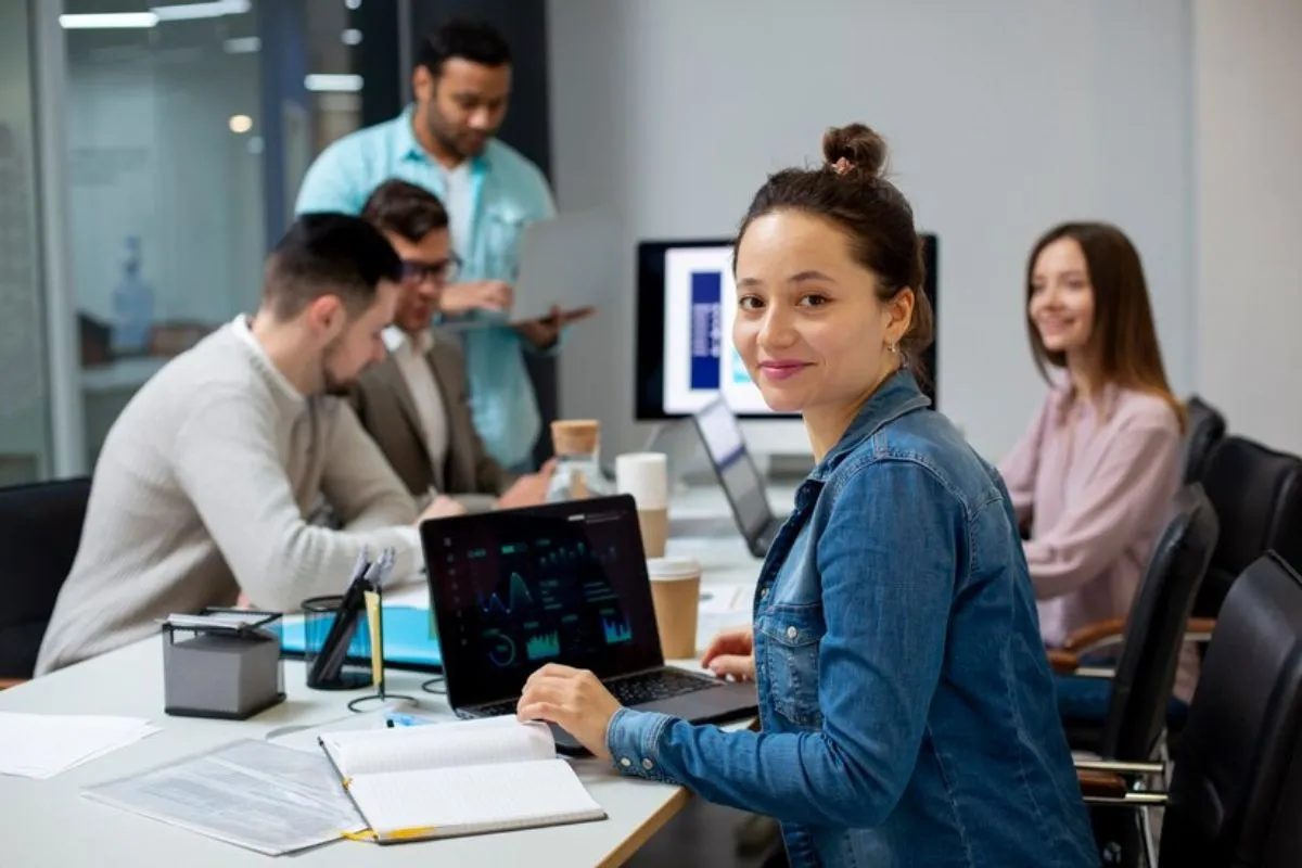 Mulher sorridente em uma reunião de negócios, trabalhando em um laptop com gráficos, ao lado de colegas em ambiente corporativo colaborativo.